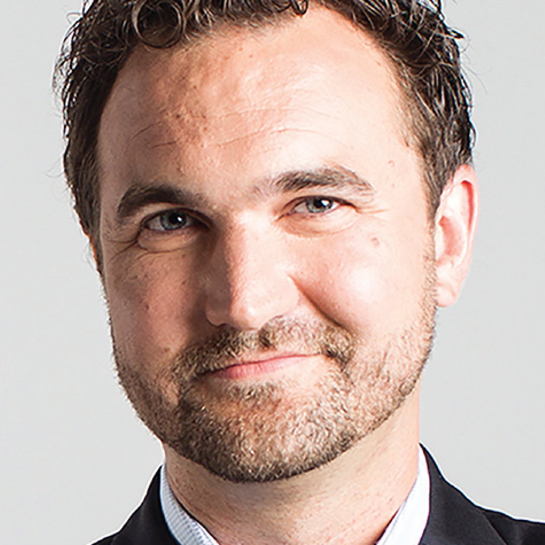 A close-up headshot of Matthew Hornsbur, smiling, with short brown hair, wearing a dark suit and light shirt.