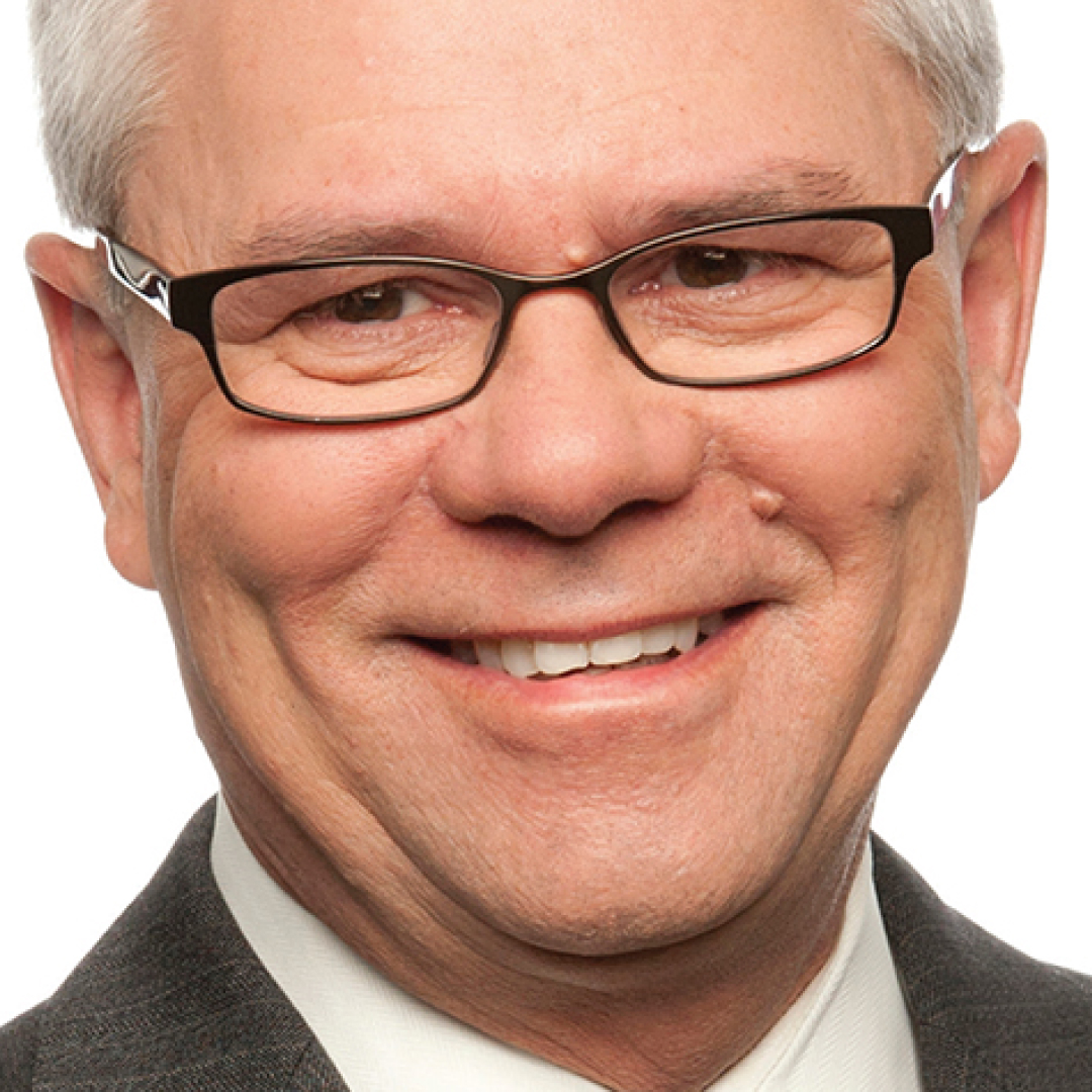 A close-up headshot of Rick Brace. He has short white hair, wears rectangular glasses, and is smiling while dressed in a dark suit and white shirt.