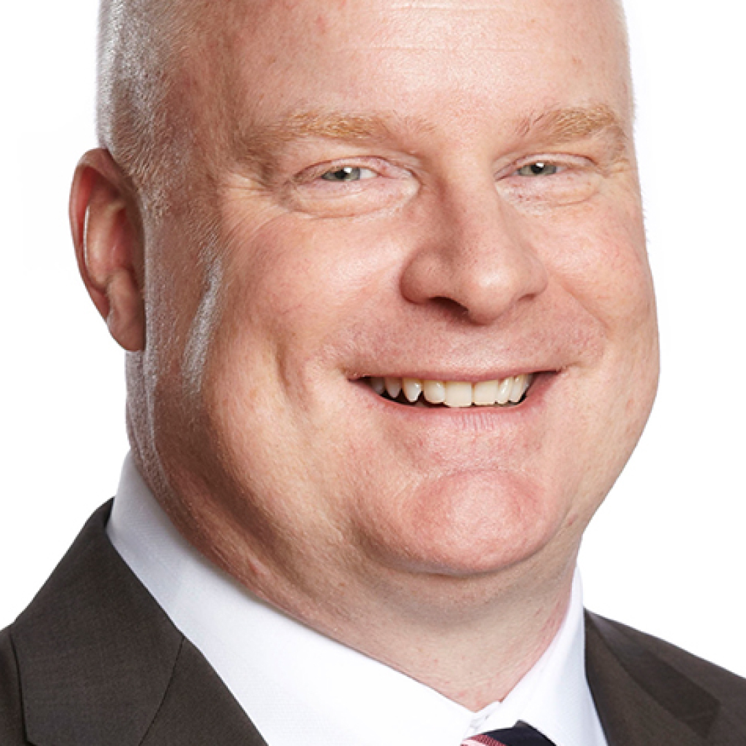 A close-up headshot of Scott Moore. He has very short hair, is smiling, and wears a dark suit, white shirt, and striped tie.
