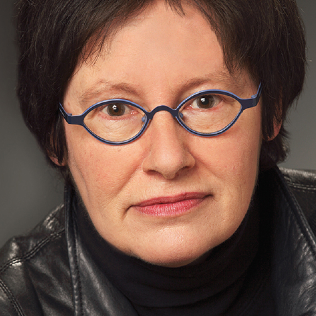 A close-up headshot of Susan Marjetti. She has short dark hair, wears round blue glasses, and has a neutral expression while wearing a dark top and jacket.
