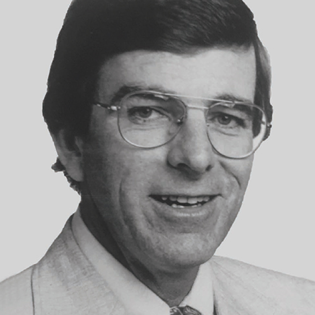 A black-and-white close-up headshot of Ted Darling. He has dark hair, glasses, and is smiling. He is wearing a light-colored suit and tie, posed against a plain backdrop.
