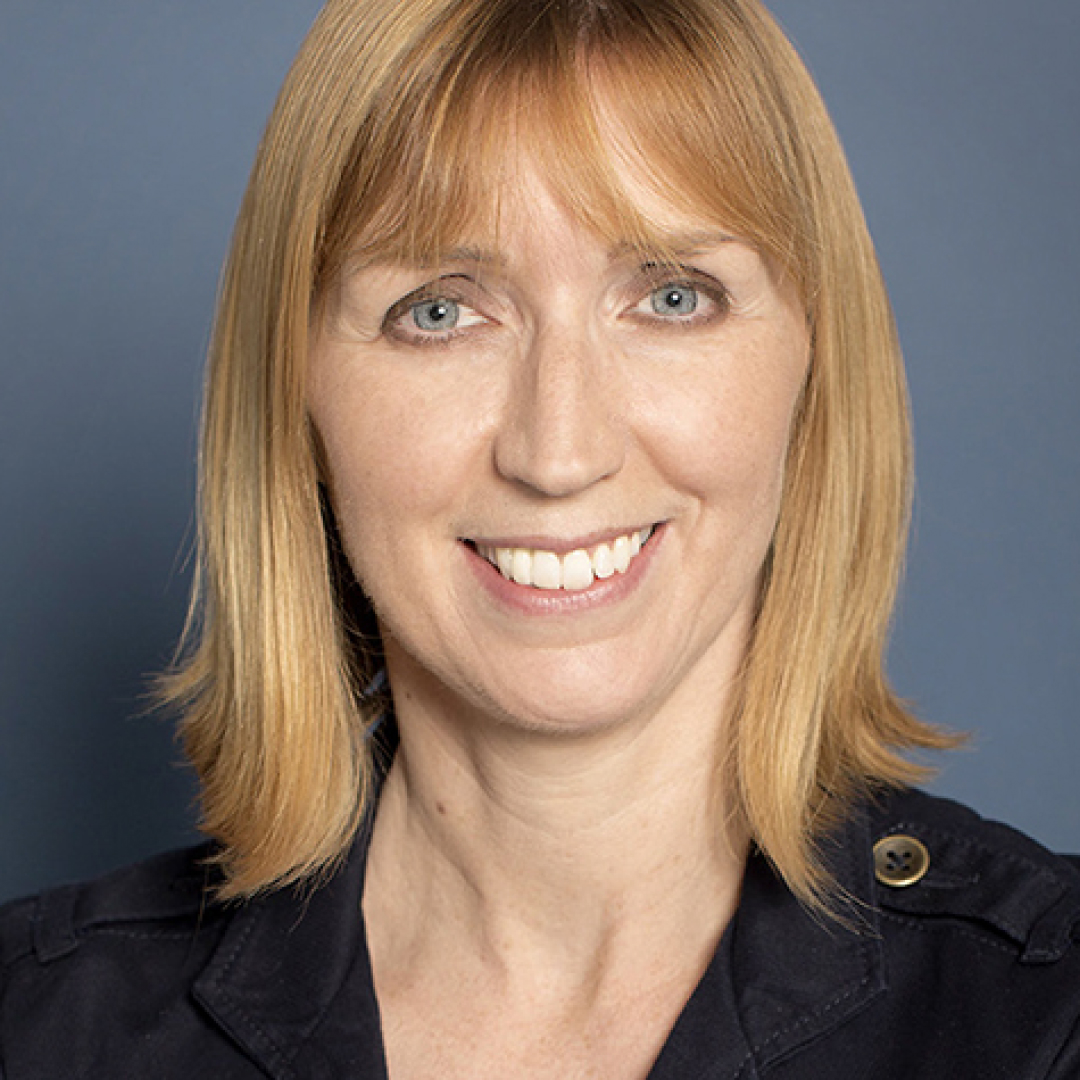 A close-up headshot of Treena Hancock. She has light blonde hair with bangs, light eyes, and is smiling. She is wearing a dark collared shirt and is posed in front of a plain blue background.
