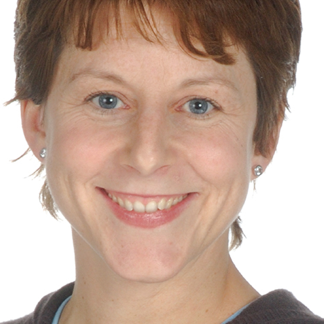 A close-up headshot of Virginia Thompson. She has short light brown hair, blue eyes, and is smiling. She is wearing stud earrings and a dark sweater over a light blue top, posed against a plain white background.