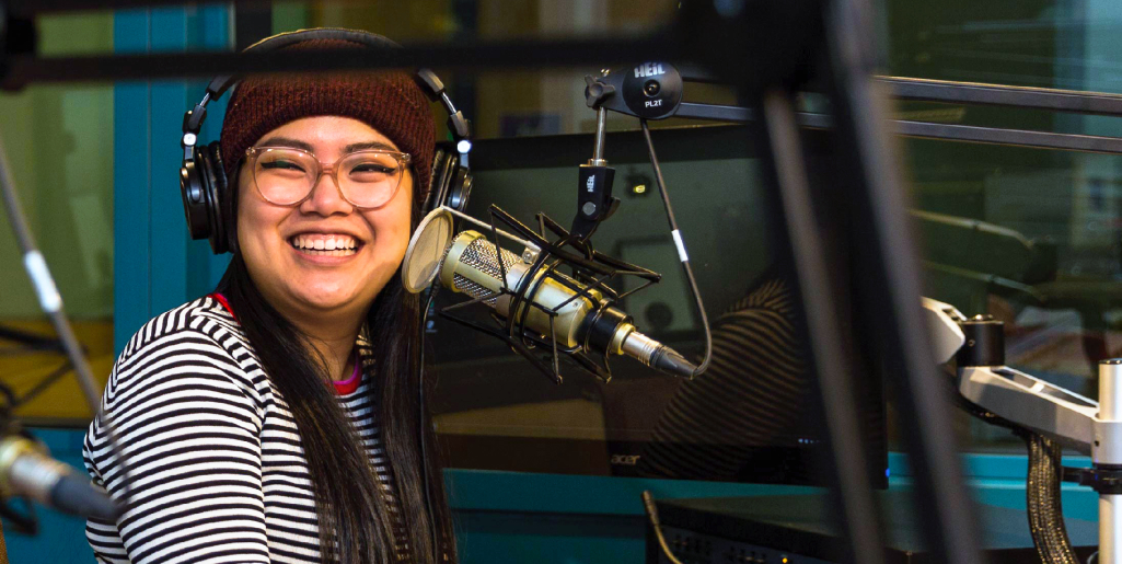 A close-up of a woman wearing glasses, headphones, and a striped shirt with a knit hat, smiling while speaking into a microphone in a radio studio.