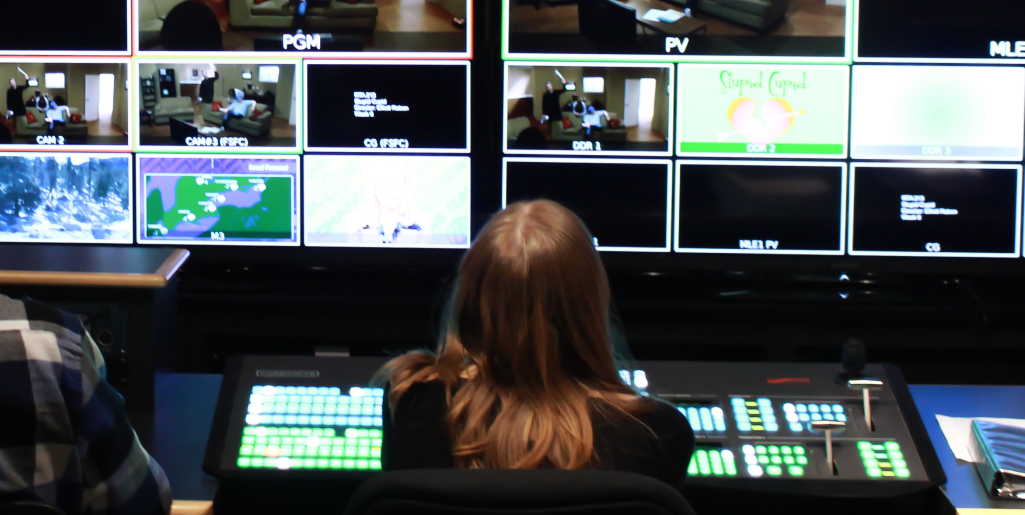 A student with long blonde hair sits at a large control panel in a television production control room, monitoring multiple screens showing different camera feeds.
