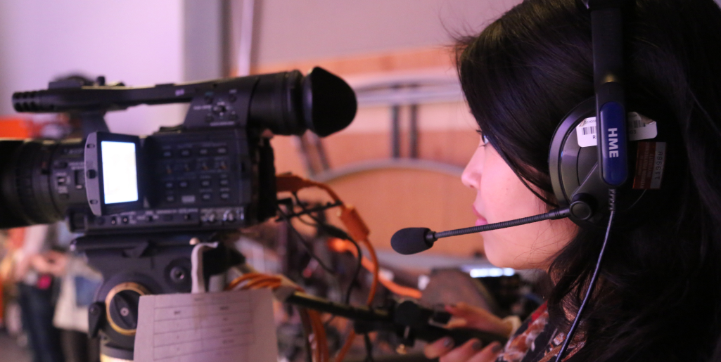 A close-up of a woman with long dark hair wearing a headset, operating a professional video camera on a tripod in a studio.