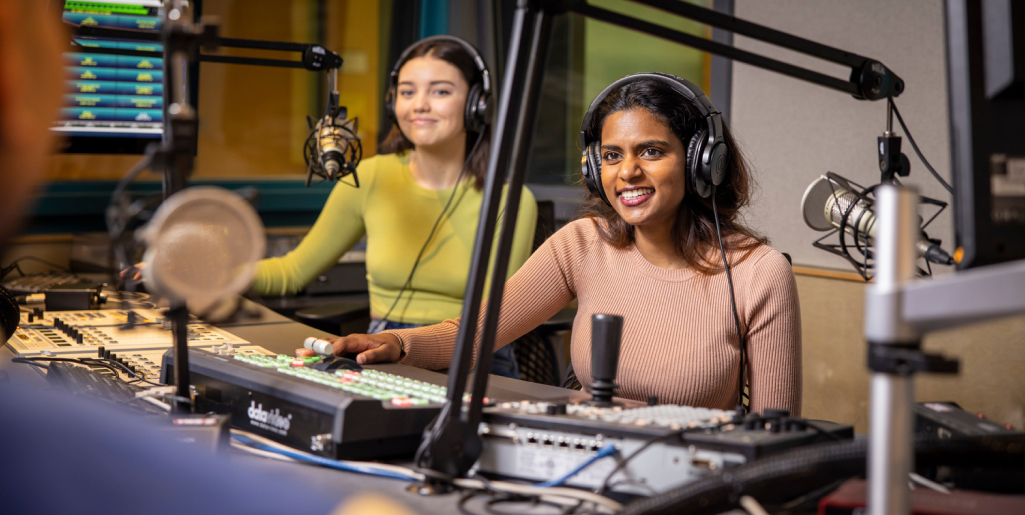 Two women wearing headphones sit in a radio studio. One, in a beige sweater, operates the soundboard and smiles, while the other, in a green top, sits beside her.