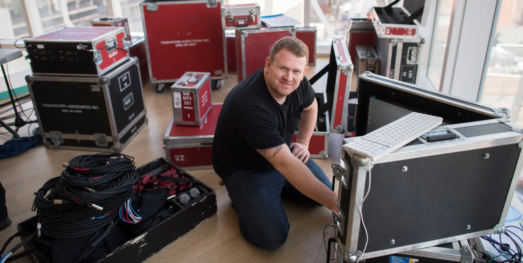 A man kneels on the floor surrounded by red and black audiovisual equipment cases, plugging cables into a large monitor box and smiling at the camera.