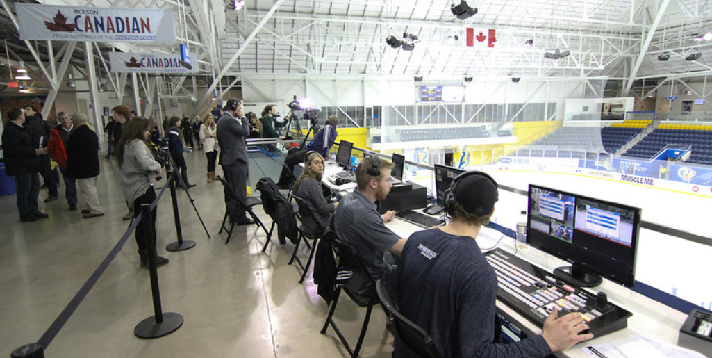 A group of students operate broadcast equipment from a booth overlooking a hockey rink, while a live game setup takes place in the background.