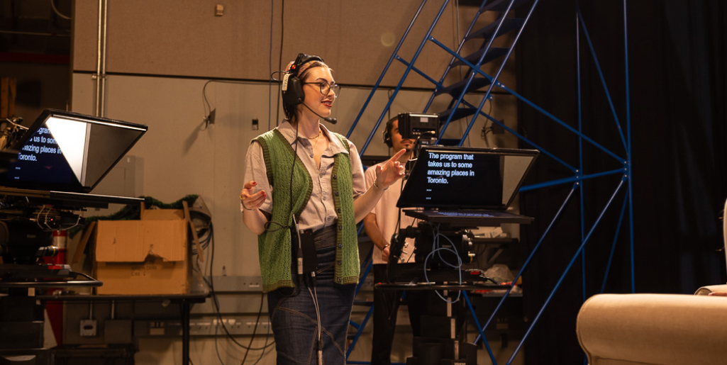 A woman wearing a headset and glasses gestures while standing in a television studio, with a teleprompter displaying text in front of her and crew members behind the camera.
