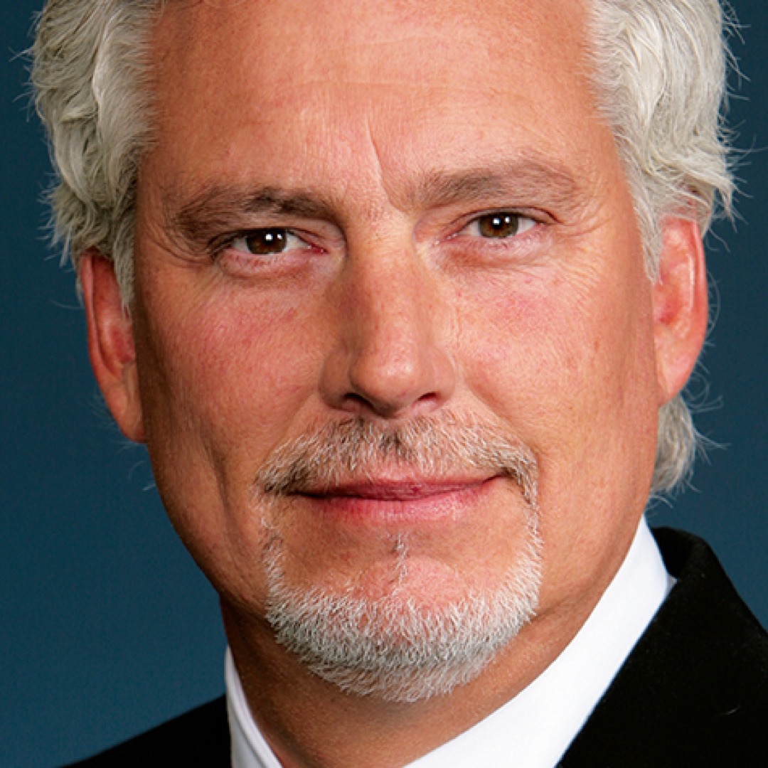 Close-up headshot of Doug Sellars with a neutral expression. He has short grey hair and a goatee and is wearing a dark suit with a white shirt.