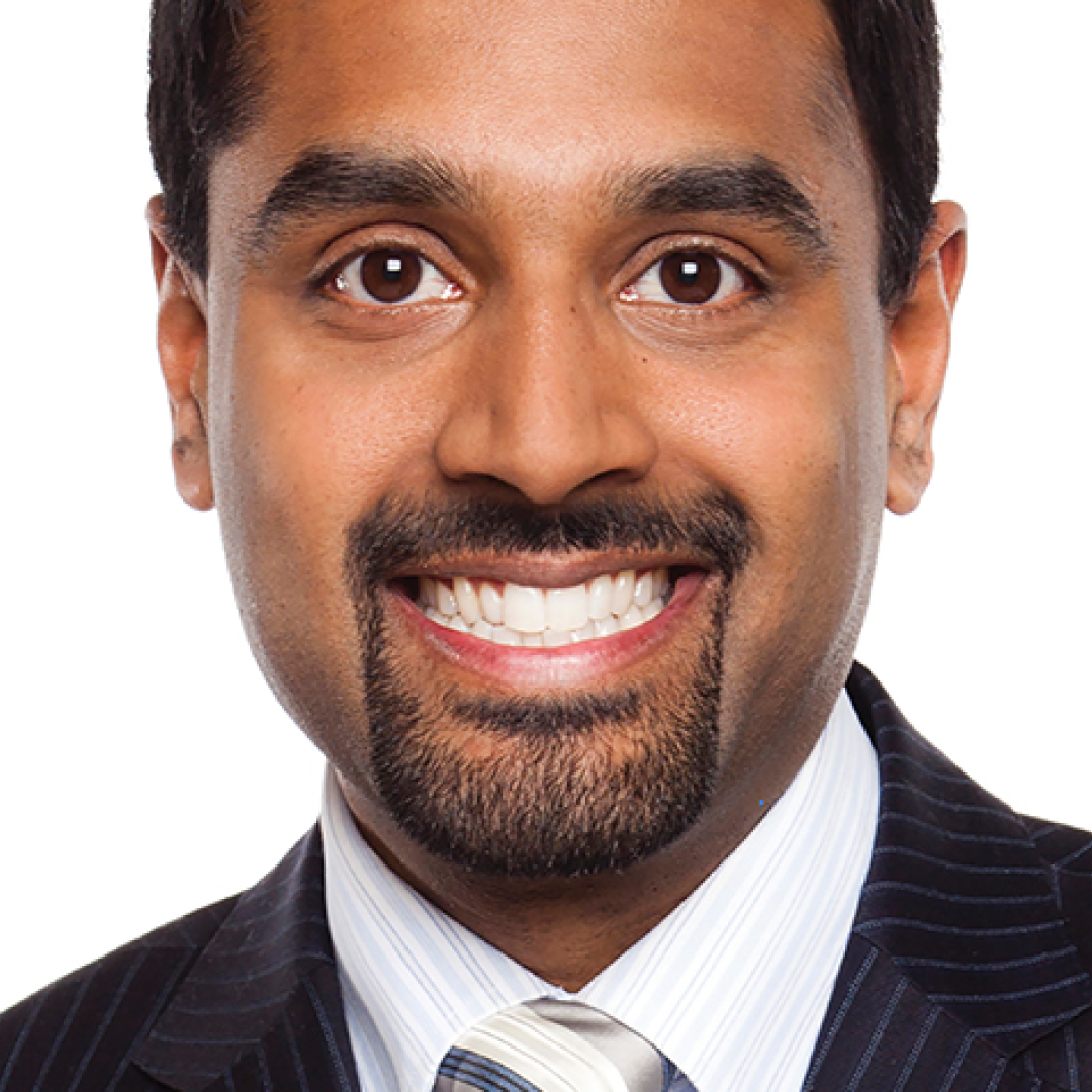 A close-up headshot of Francis D’Souza. He has short black hair, a goatee, and is smiling widely. He is wearing a pinstripe suit with a white shirt and tie, posed against a plain white background.