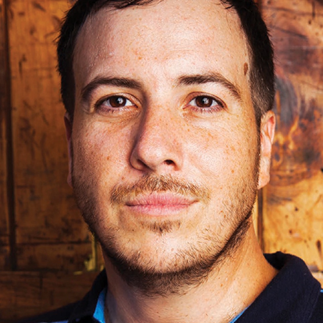 Close-up headshot of JJ Johnson smiling. He has short dark hair and light facial hair and is wearing a striped collared shirt.