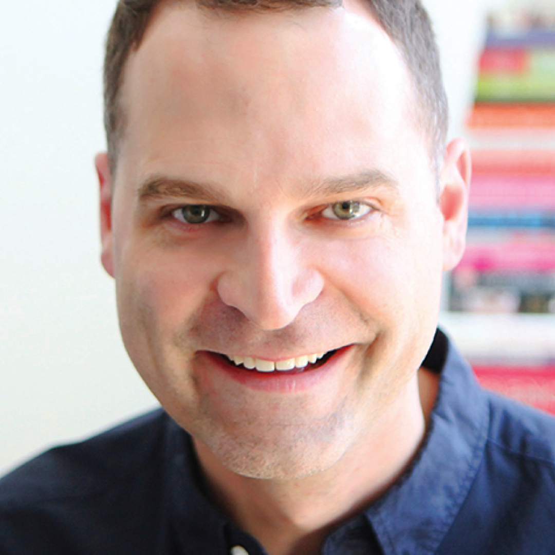 Close-up headshot of Jay Onrait smiling. He has short hair and is wearing a dark collared shirt.