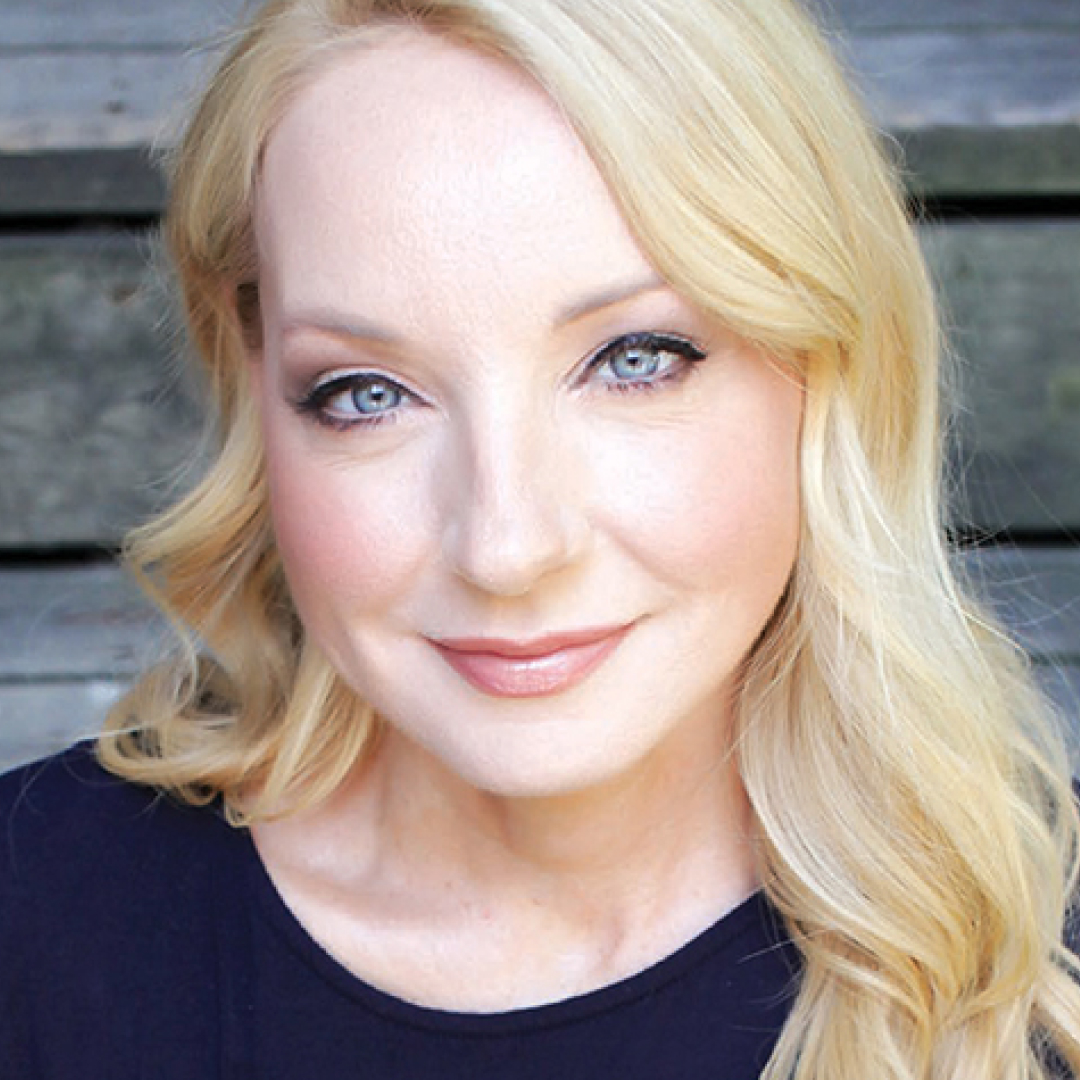 Close-up headshot of Jessica Holmes smiling. She has long blonde hair and is wearing a dark top.