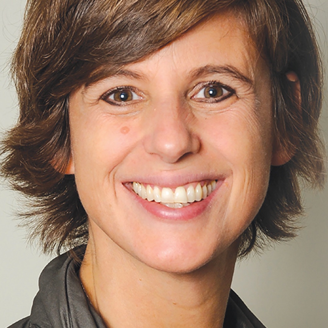 Close-up headshot of Julie Adam smiling. She has short brown hair and is wearing a dark jacket.