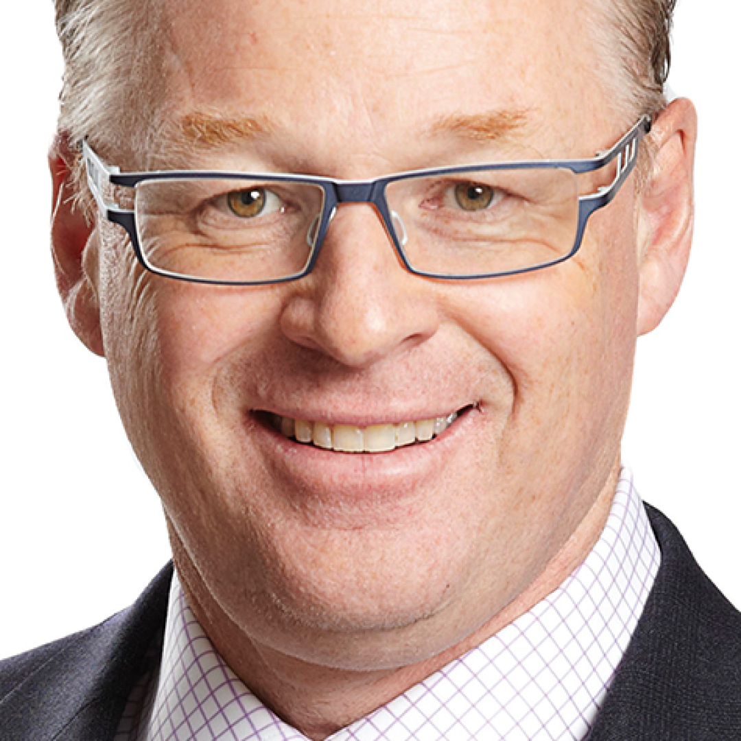 Close-up headshot of Keith Pelley smiling. He has short grey hair and glasses and is wearing a suit with a checkered shirt and tie.