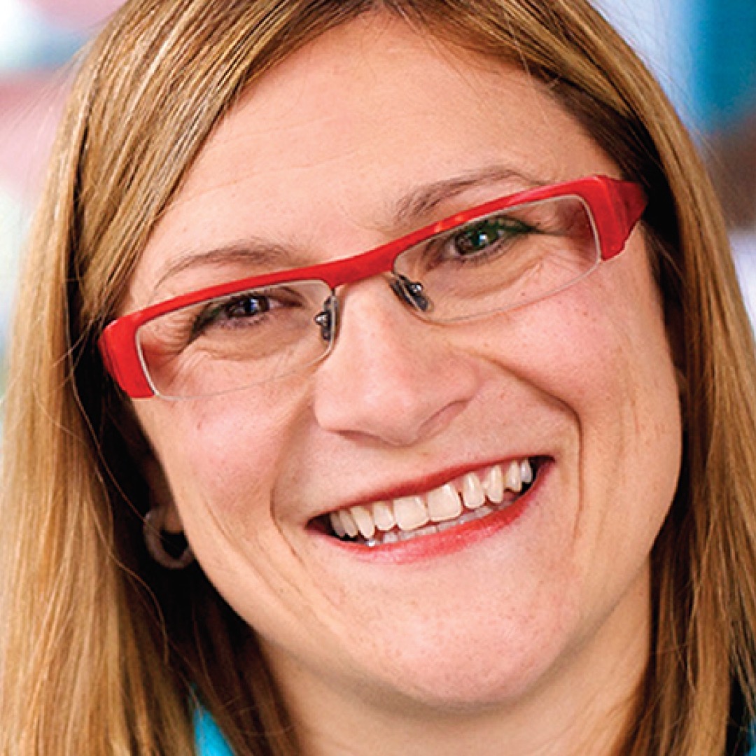 A close-up headshot of Kim Wilson, smiling, with straight light brown hair, wearing red glasses and a blue top.