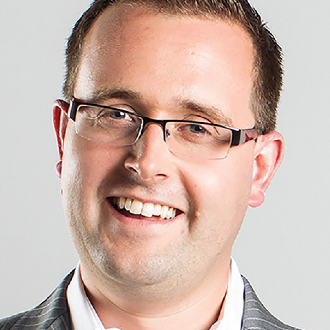 A close-up headshot of Mark Bishop, smiling, with short brown hair, wearing glasses, a striped jacket, and a white shirt.
