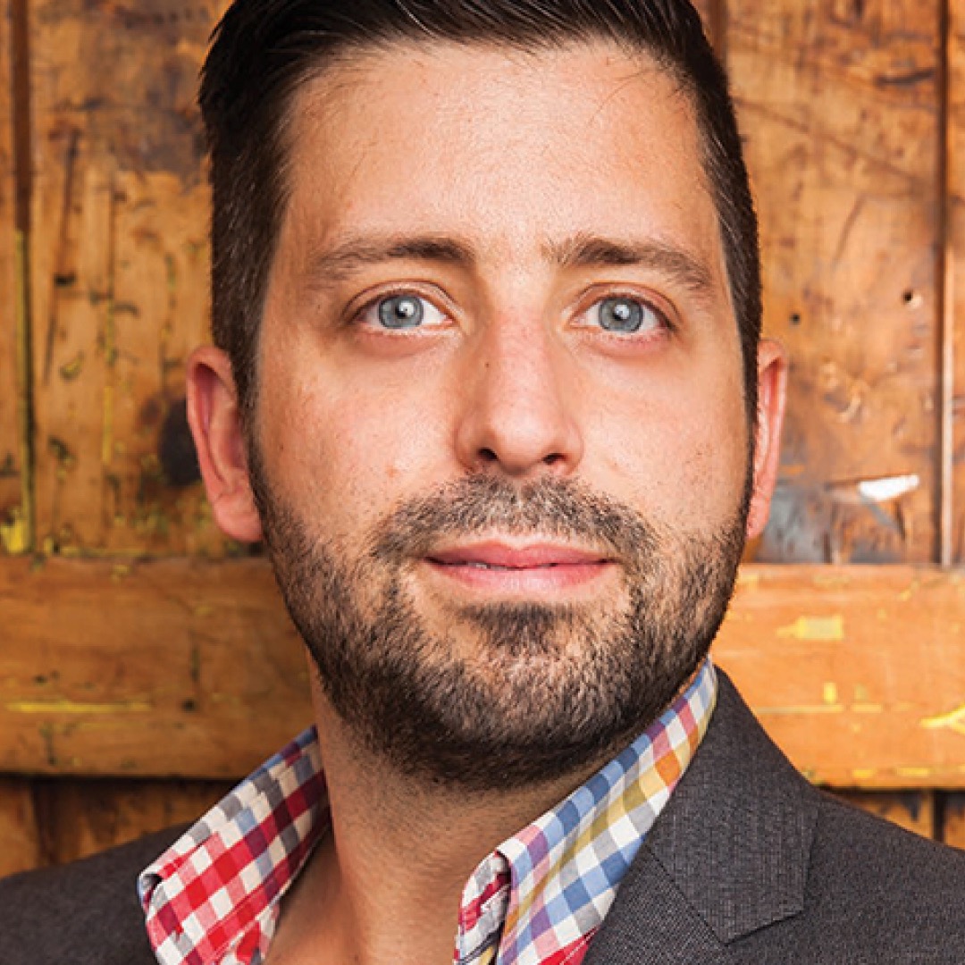 A close-up headshot of Matt Bishop, with short dark hair and a trimmed beard, wearing a checkered shirt and dark suit jacket.
