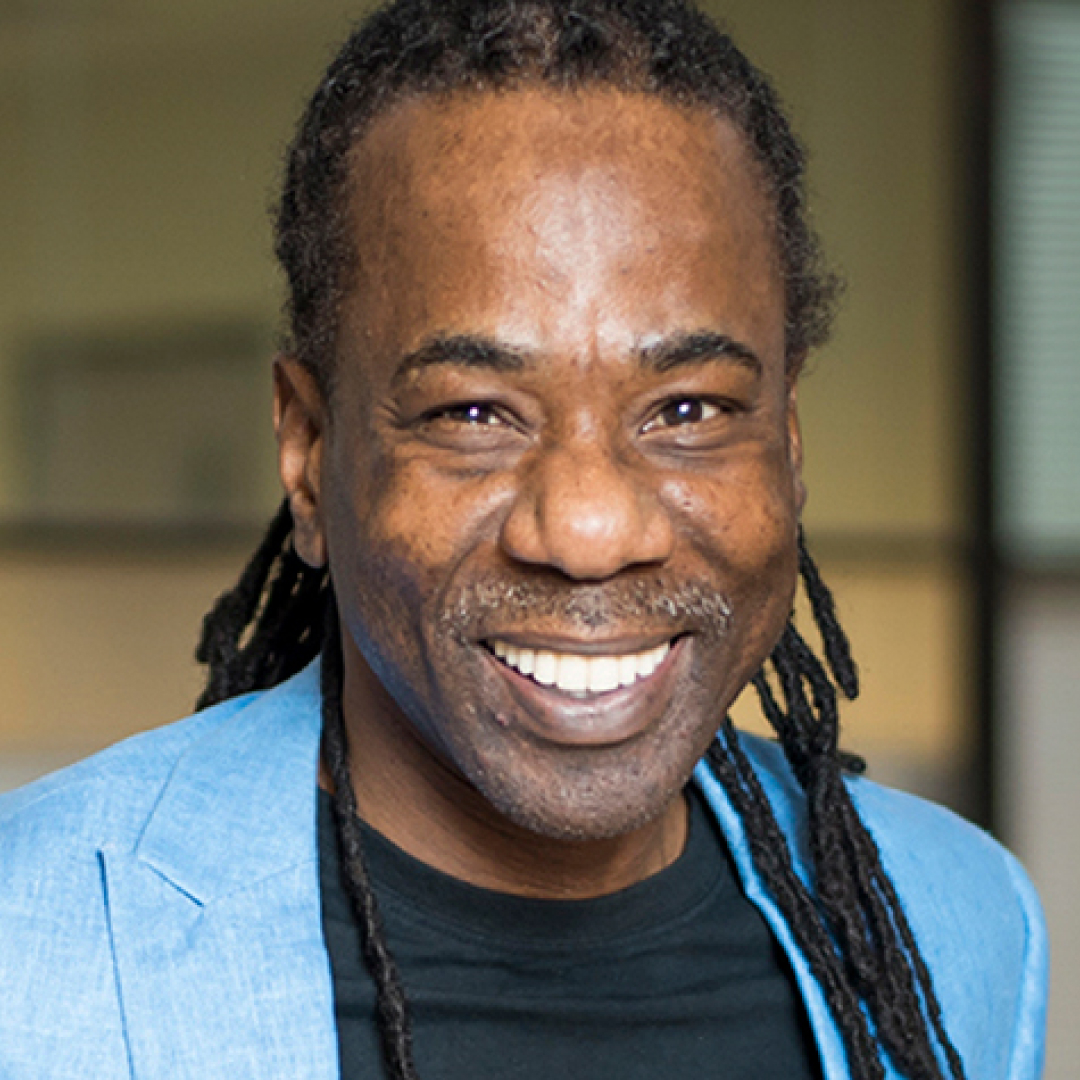 A close-up headshot of Ron Nelson. He has long braided hair, a mustache, and goatee, and is smiling widely while wearing a light blue blazer over a black shirt.