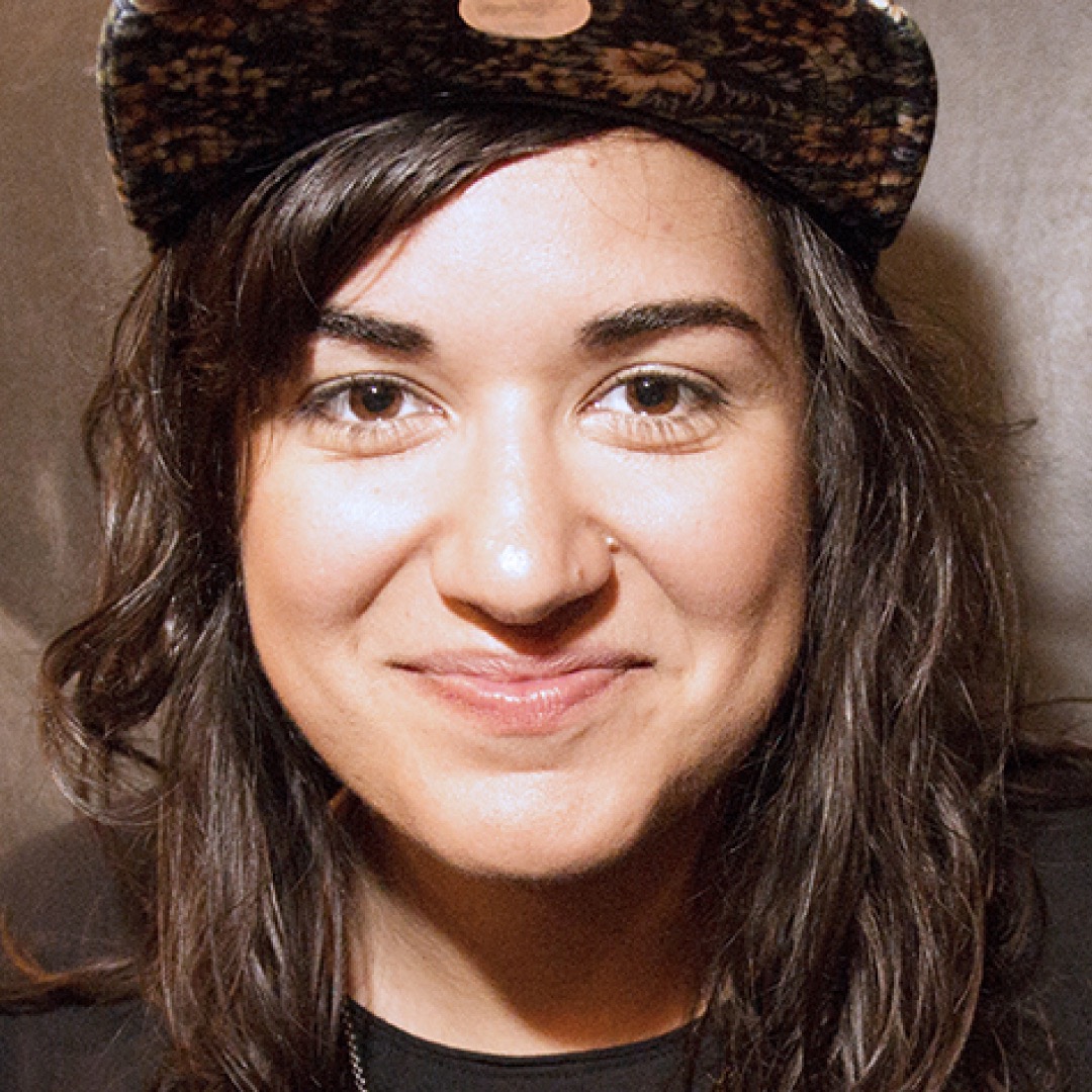 A close-up headshot of Sabrina Jaless. She has long wavy dark hair, a nose piercing, and is smiling while wearing a patterned cap.