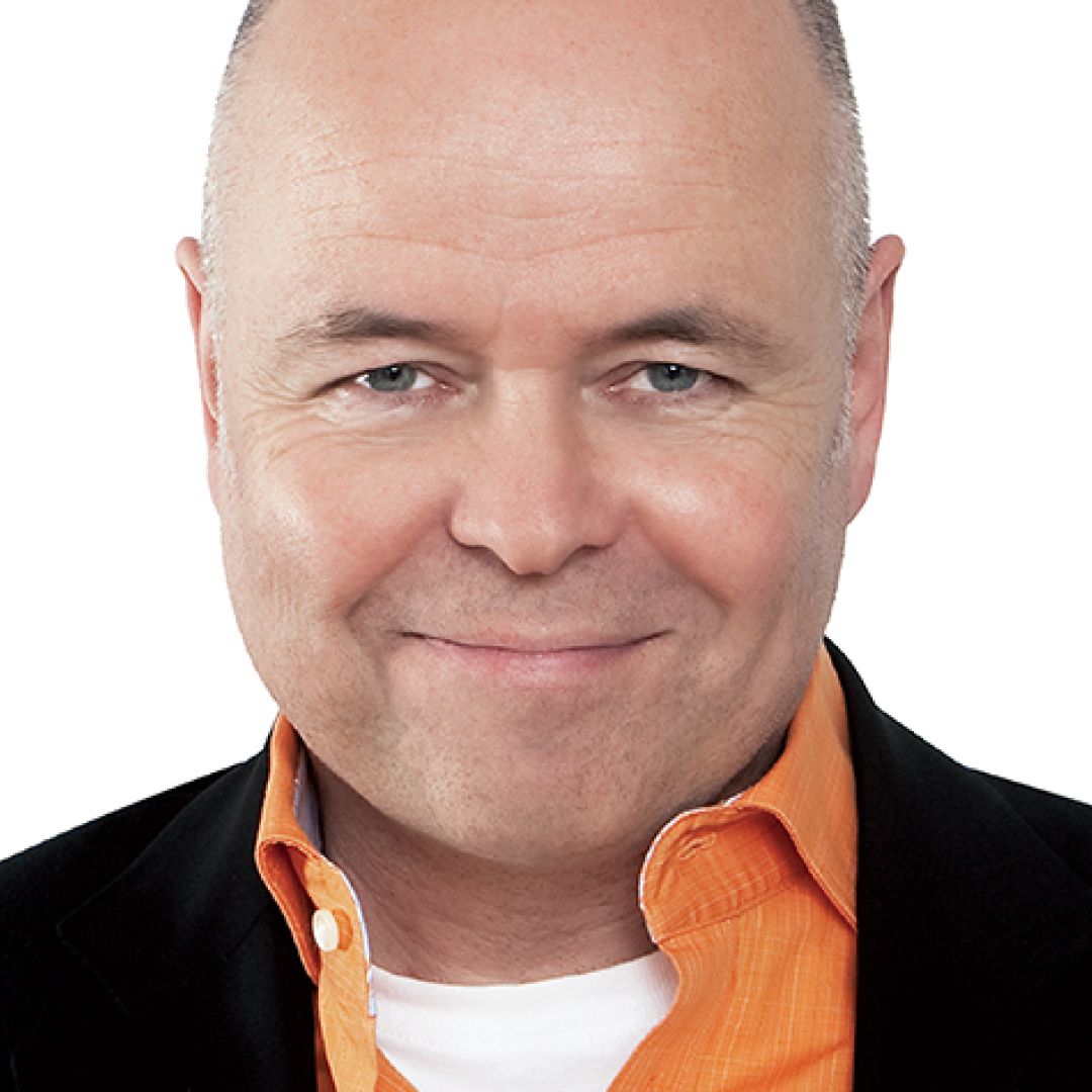 A close-up headshot of Terry O’Reilly. He has a shaved head, wears a black jacket with an orange collared shirt, and is smiling with a confident expression.