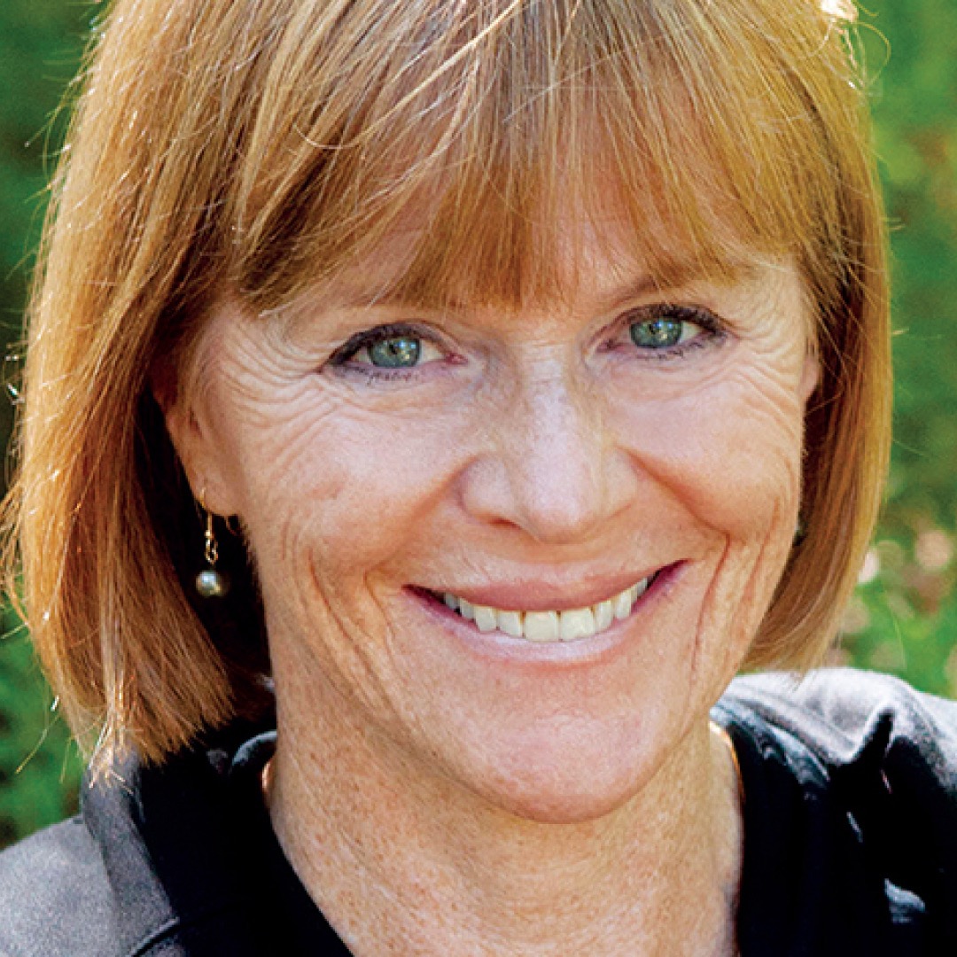 A close-up headshot of Valerie Pringle. She has light red hair with bangs, light eyes, and is smiling. She is outdoors with greenery blurred in the background, wearing a dark top and drop earrings.