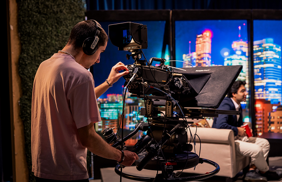 A medium shot of a man operating a large recording camera.  In the background, you can see a talk shsow setting and a man in a navy blue suit sitting on an armchair and speaking to someone off screen.