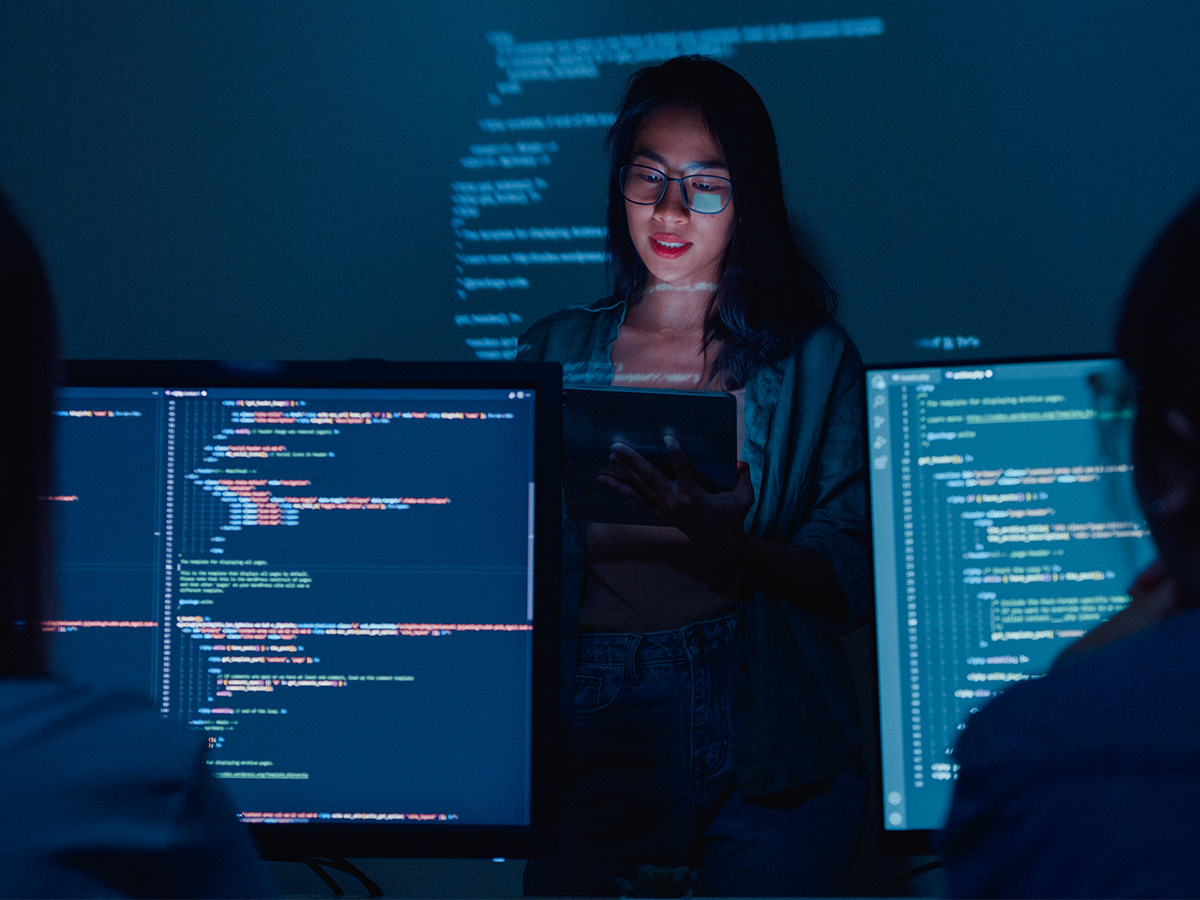 Student holding a tablet in a dark room with code projected in the background, and two monitors with code in the foreground