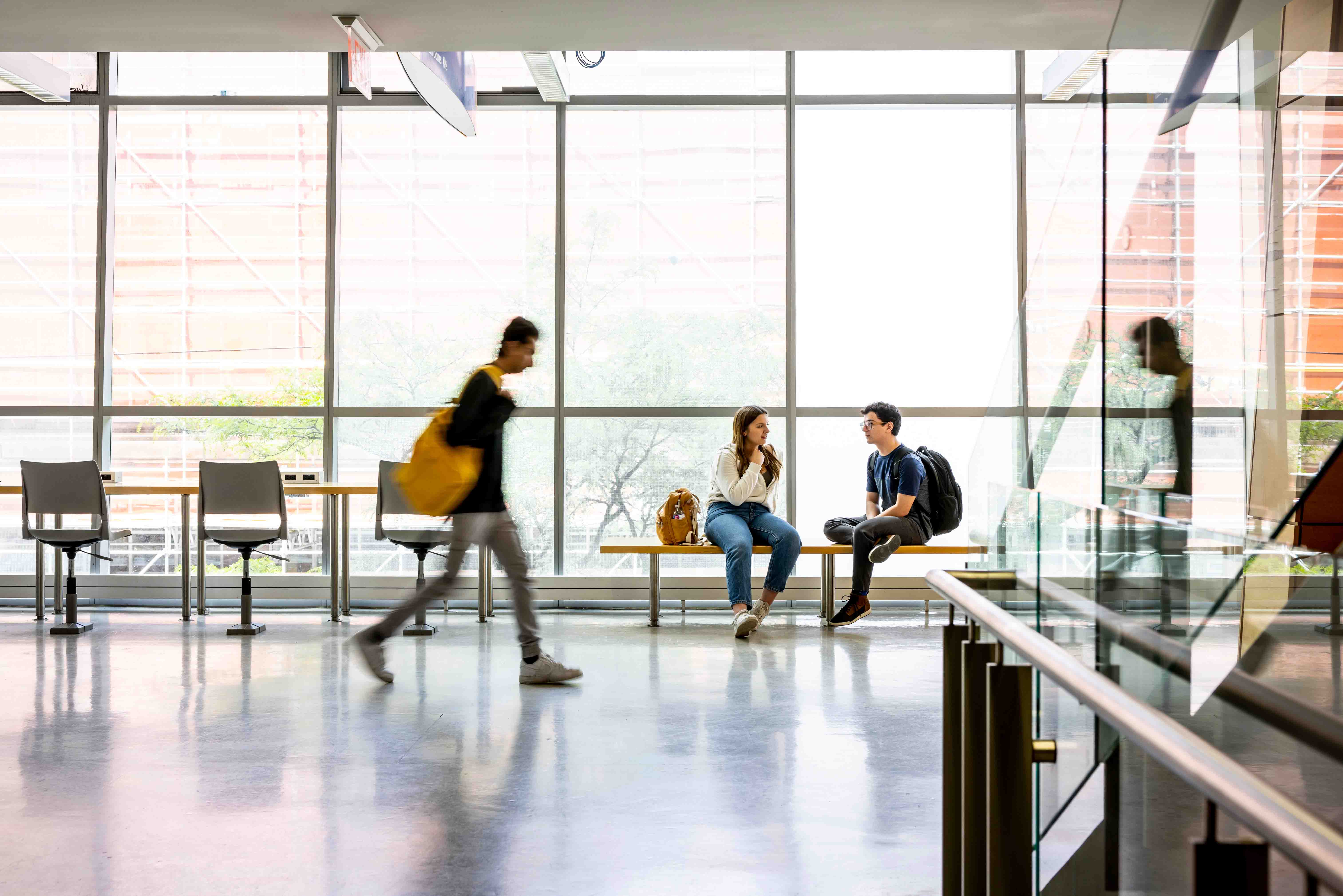 Students sitting and talking in a campus hallway while another student walks by.