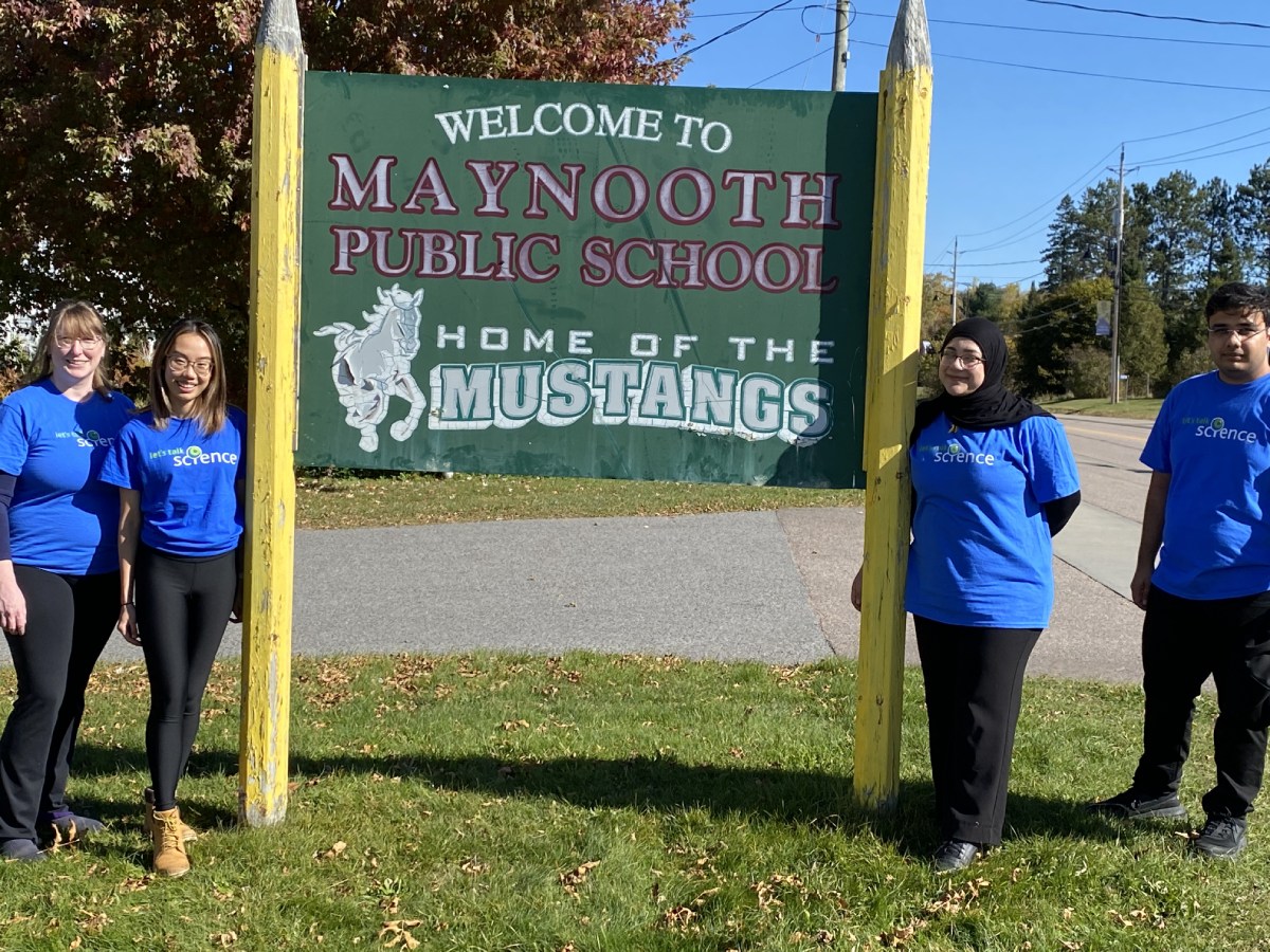 SciXchange staff and volunteers standing outside infront of a school sign that reads: "Welcome to Maynooth Public School, Home of the Mustangs" a symbol of a mustang horse is shown. 
