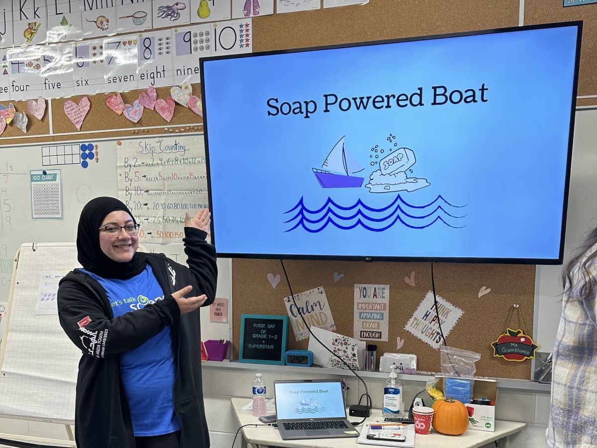 Zayneb Al-Hantoshi stands infront of a classroom monitor that reads: "Soap Powered Boat" with an illustration of a boat and a soap bar, on top of waves. 