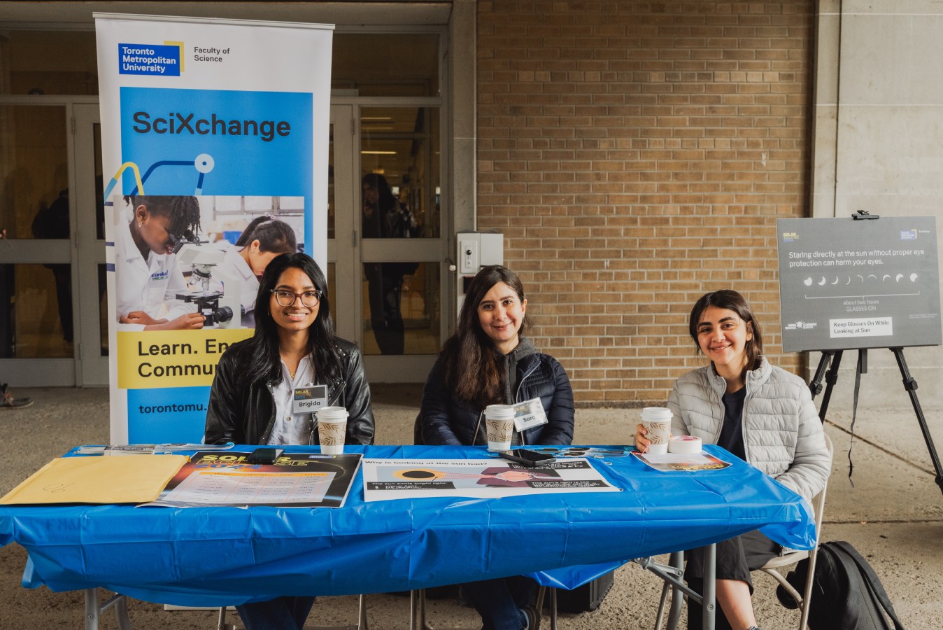 Three volunteers sit at a stand at the SciXchange event. Graphics and illustrations are displayed on the table