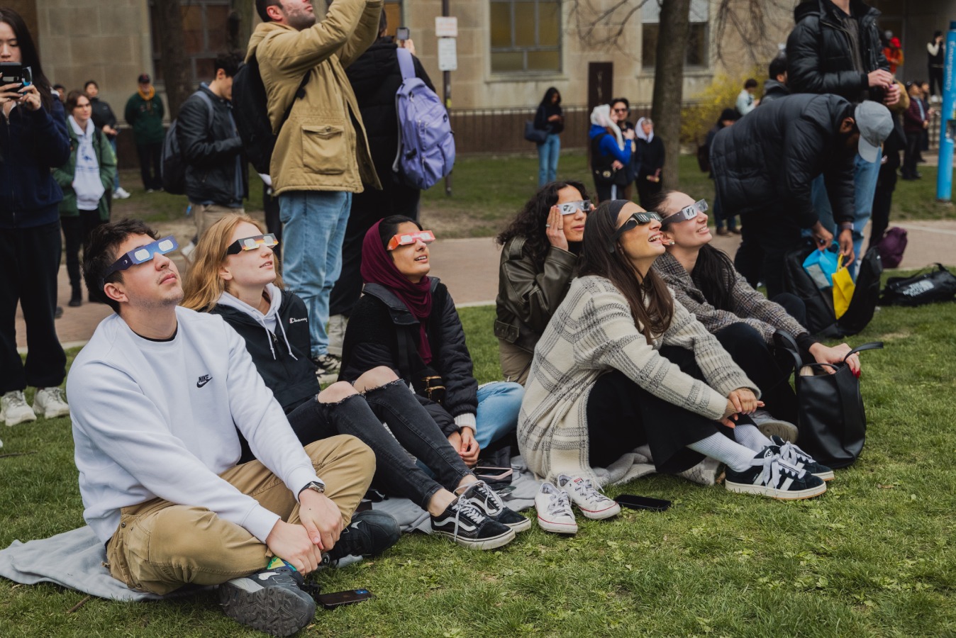 Reeda Mahmood and six other classmates sit on the grass outside TMU community park, looking up at the sky while wearing eclipse glasses