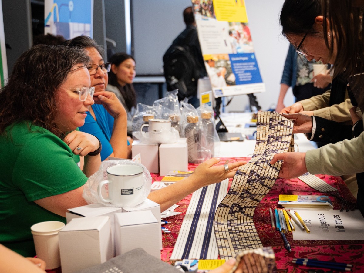 Brooke Filsinger, shows a wampum belt to a guest who is inspecting it. Brooke Filsinger and others are sitting down at a table with various merchandise and items displayed on the table