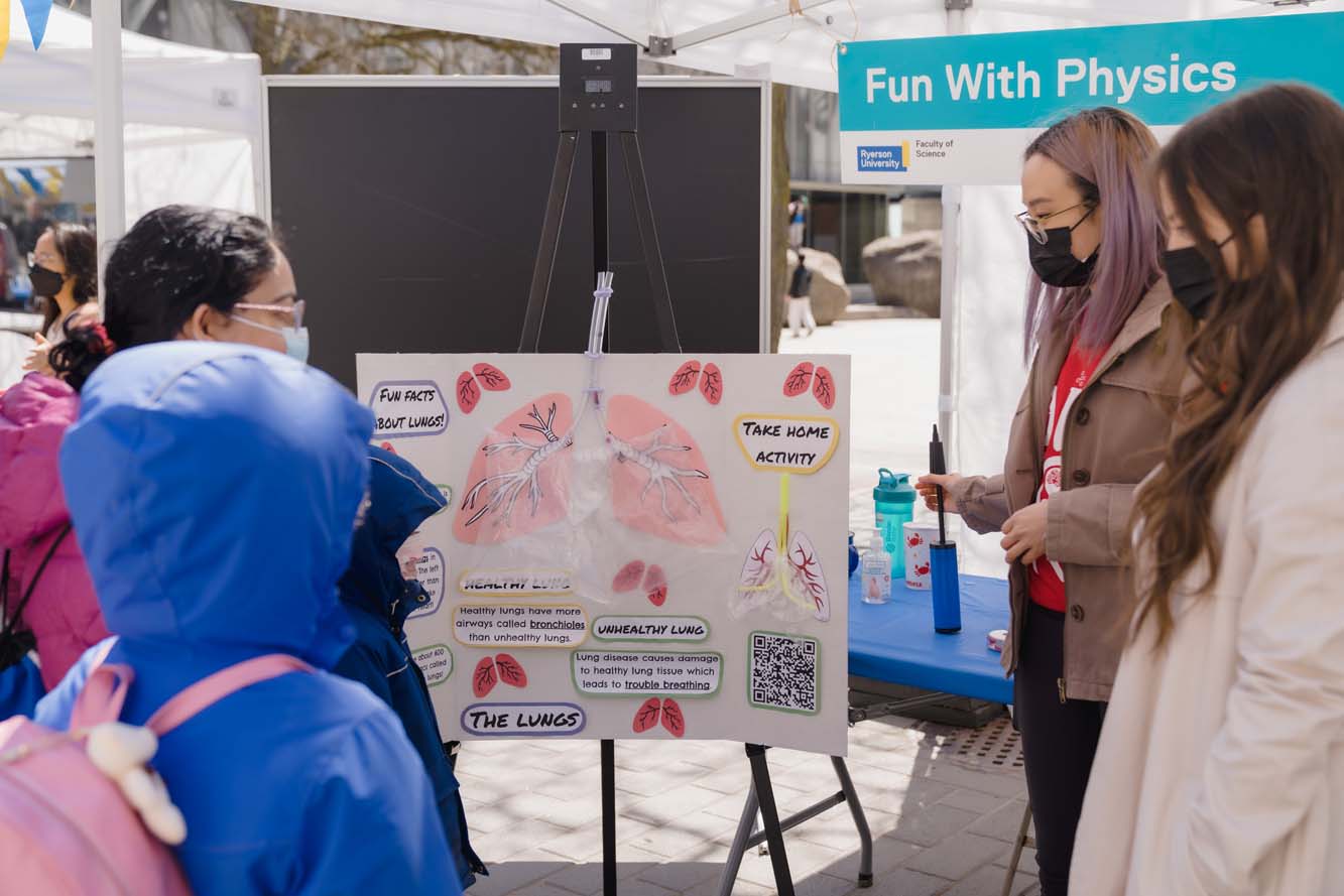Two volunteers demonstrating a presentation board with information about the lungs to a few children and adults at the Fun With Physics booth.