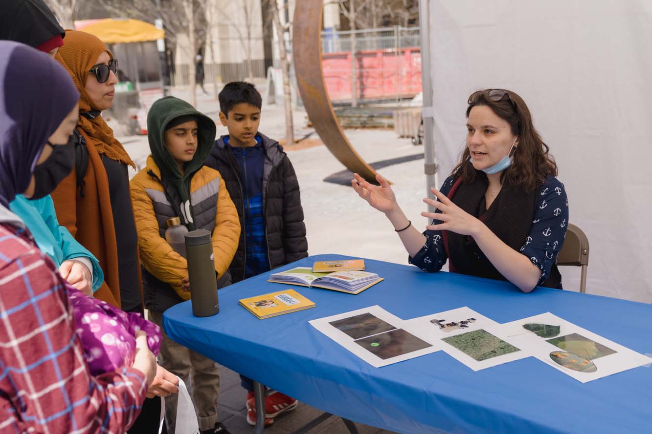 A volunteer talking to a group of children and adults at a booth.