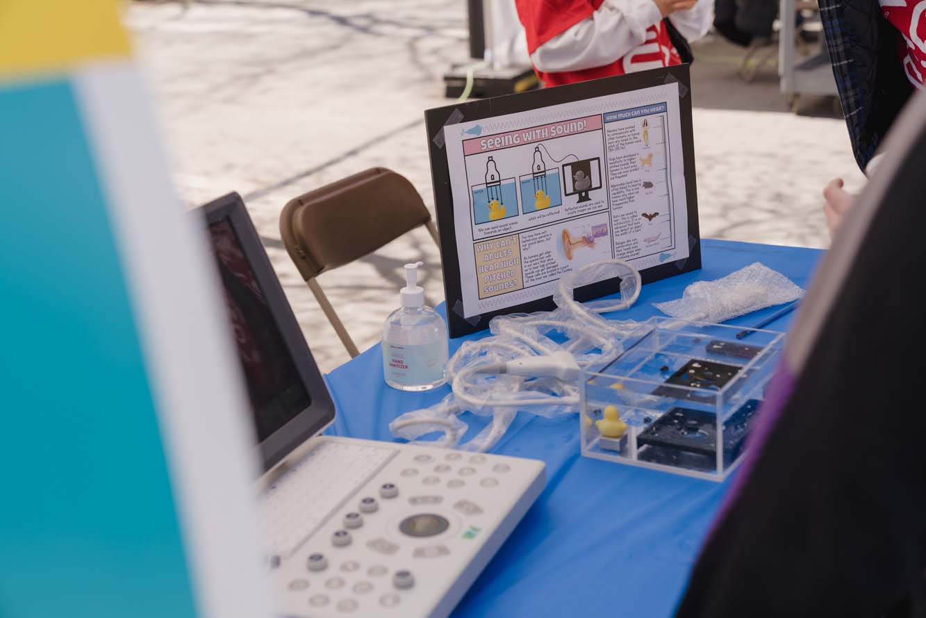 An ultrasound machine, a rubber duck and a black device in a transparent container filled with water and a presentation board about "Seeing with Sound" on the table of a booth.