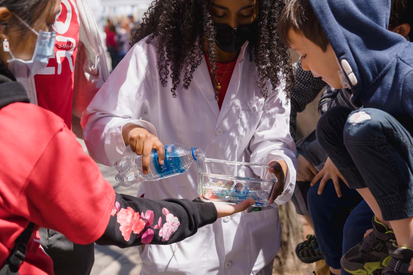 A volunteer pouring water into a glass container containing some food colouring as some children watch.