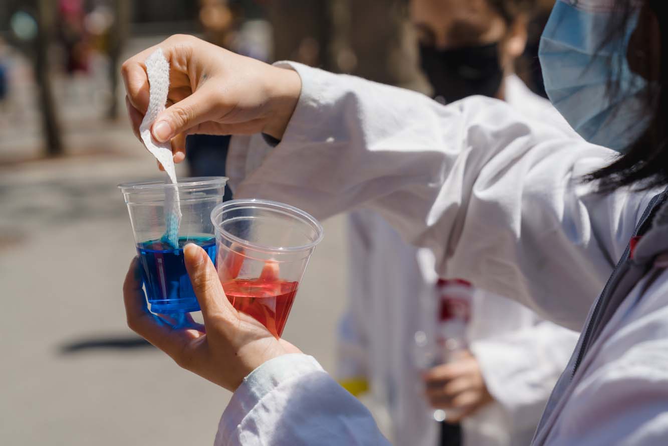 A volunteer wearing a white lab coat dipping a piece of paper towel into a cup filled with blue liquid.