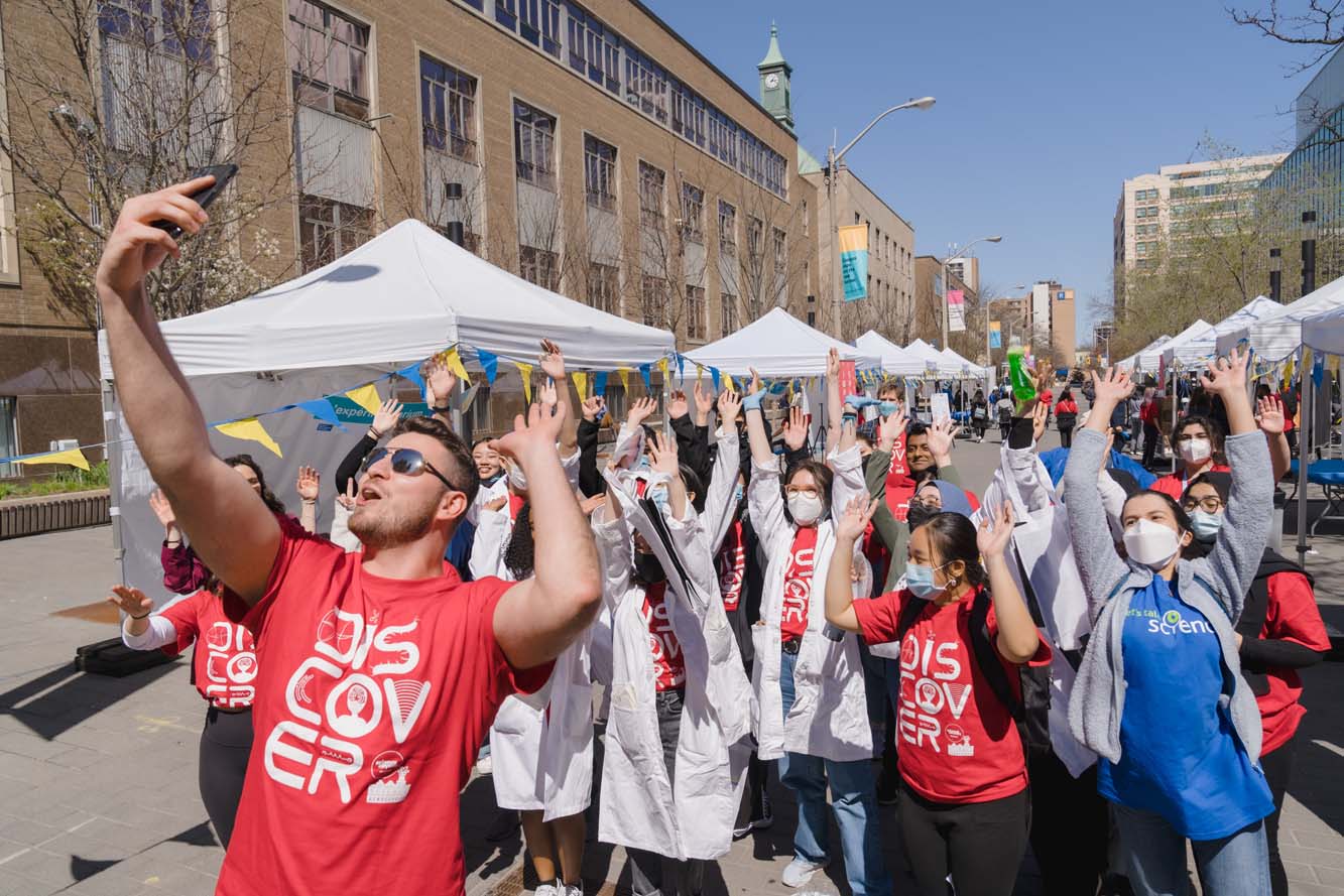 Group of about 30 people, many wearing red t-shirts, some wearing white lab coats, raising their hands and posing for fun, with white event tents in the background.