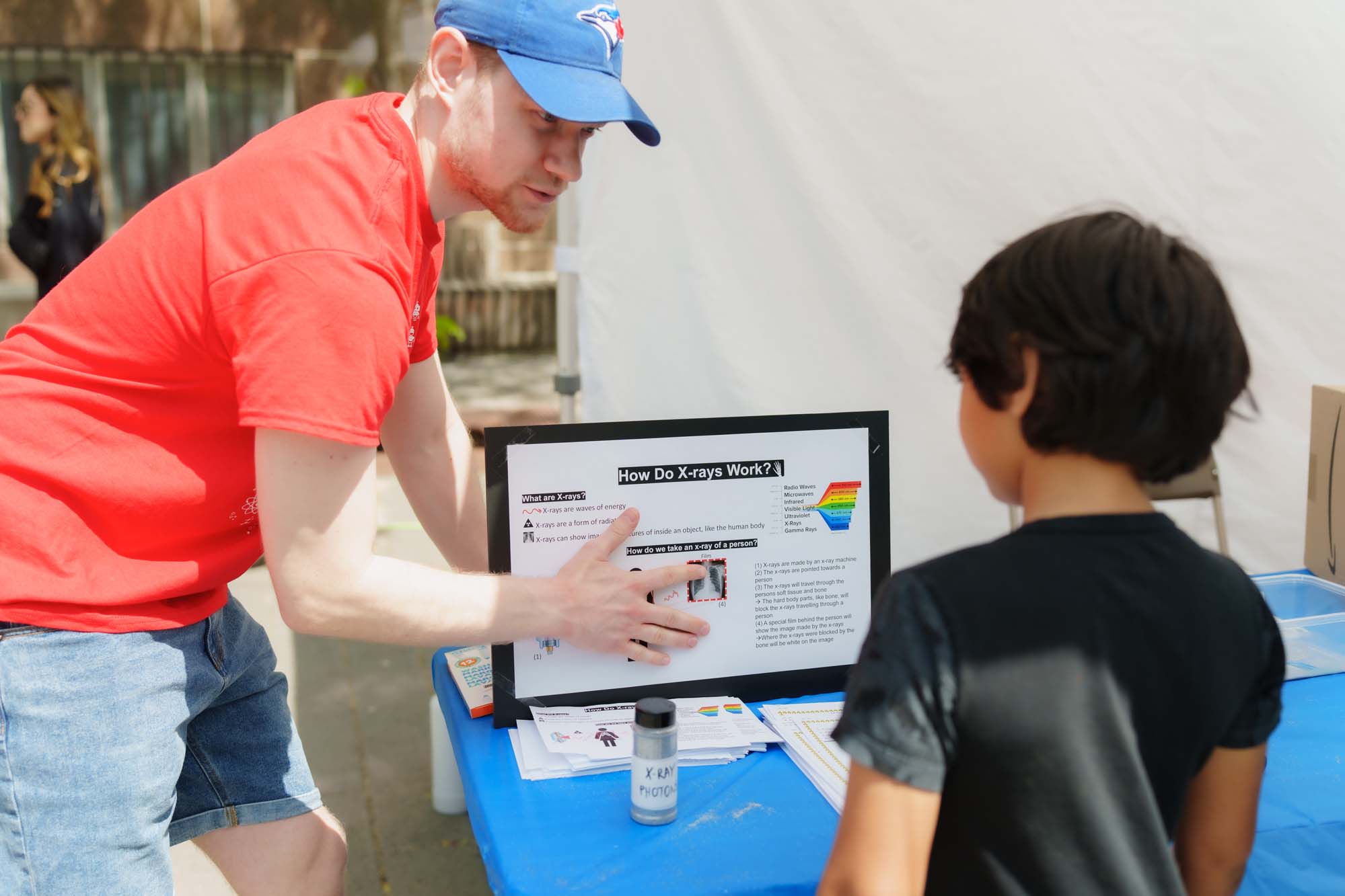 A volunteer pointing at a presentation board about "How Do X-rays Work?" to a child.