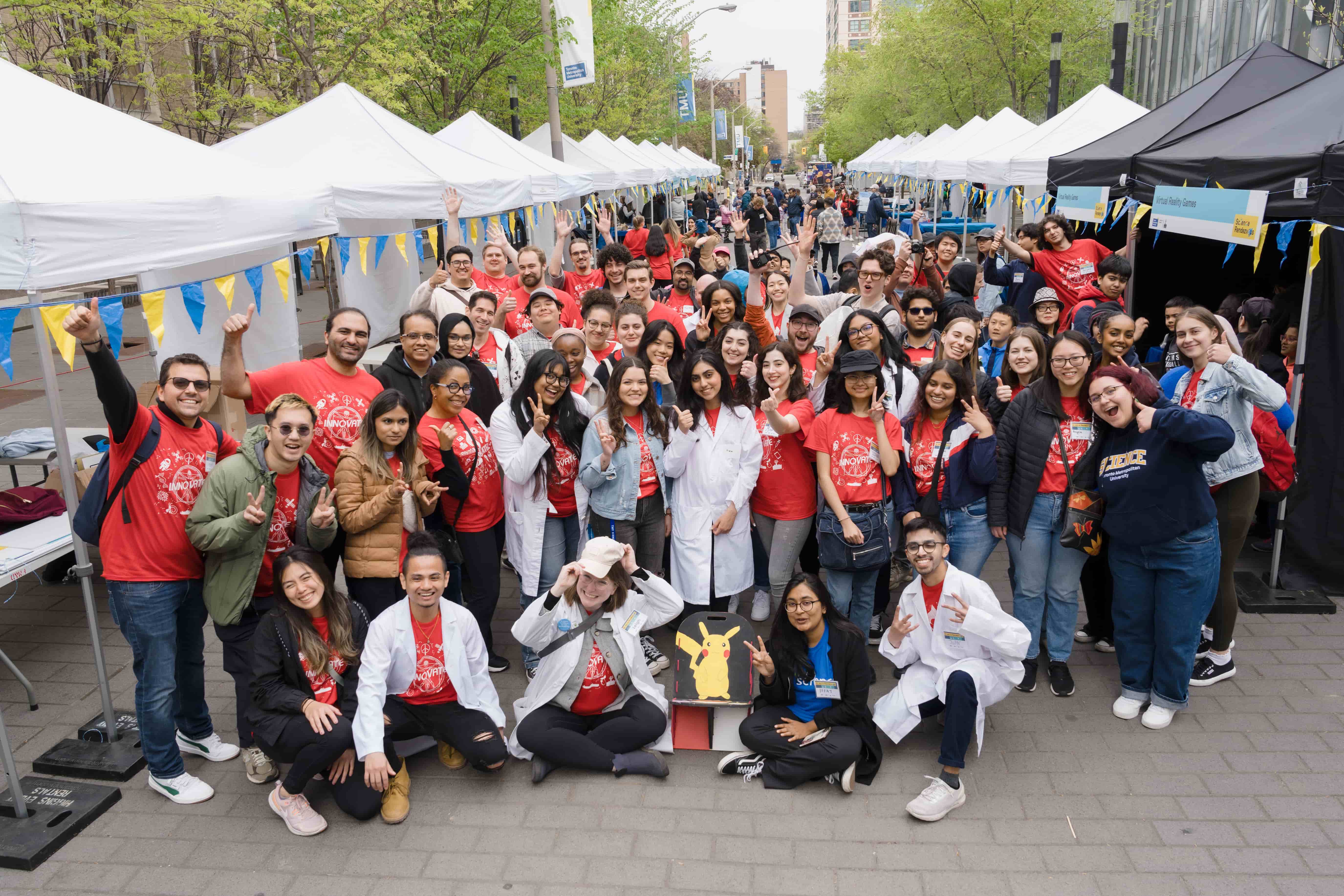 Group of about 40 people, wearing red t-shirts, posing for fun, with white event tents in the background.