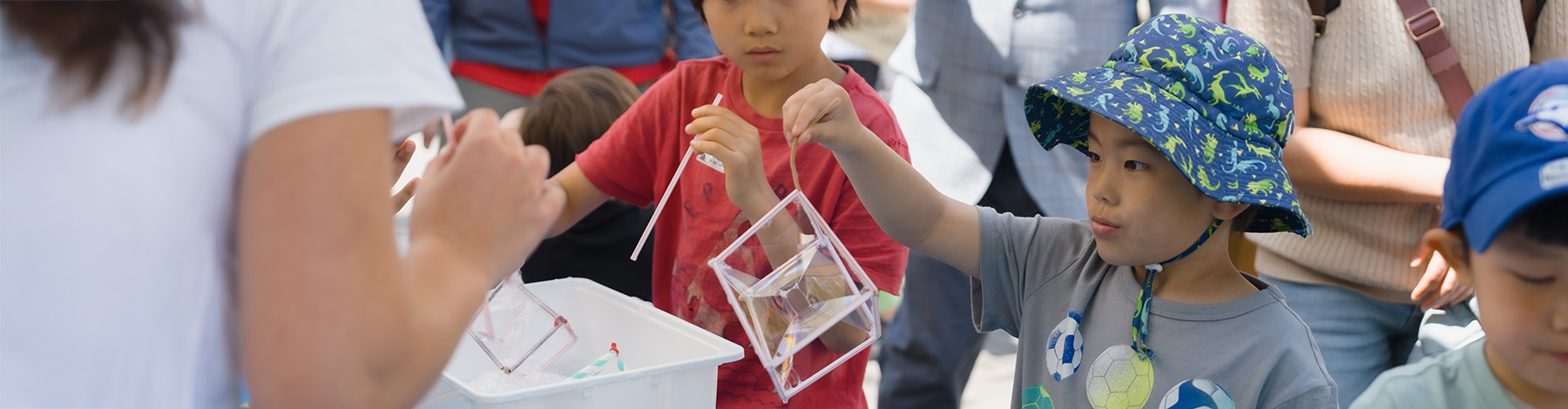 Children participate in a hands-on activity outdoors, holding clear cube-shaped containers and using straws, while an adult guides them.
