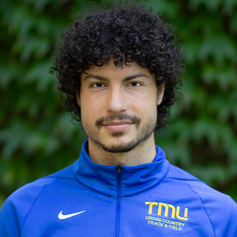 Headshot of Aaron McQuaid with dark, curly hair, beard, with soft smile, wearing blue TMU jersey with greenery in background