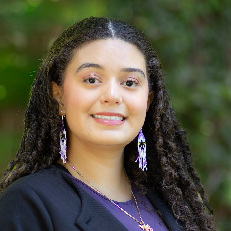 Headshot of young Indigenous woman with curly dark brown hair, smiling showing teeth, wearing beaded drop earrings against blurred green background