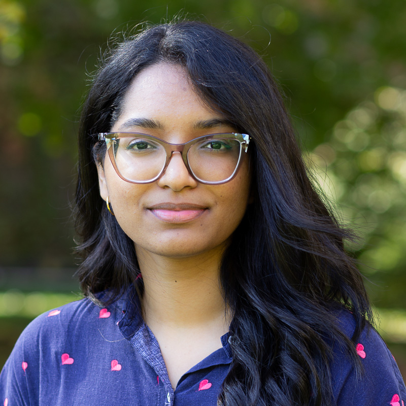 Headshot of Brigida, with long dark hark, wearing glasses, smiling gently, wearing blue shirt 