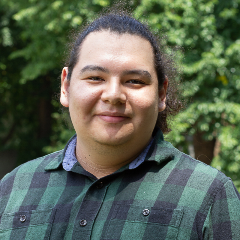 Headshot of Caleb Wesley. Caleb is wearing a green plaid shirt with light green foliage in the background.