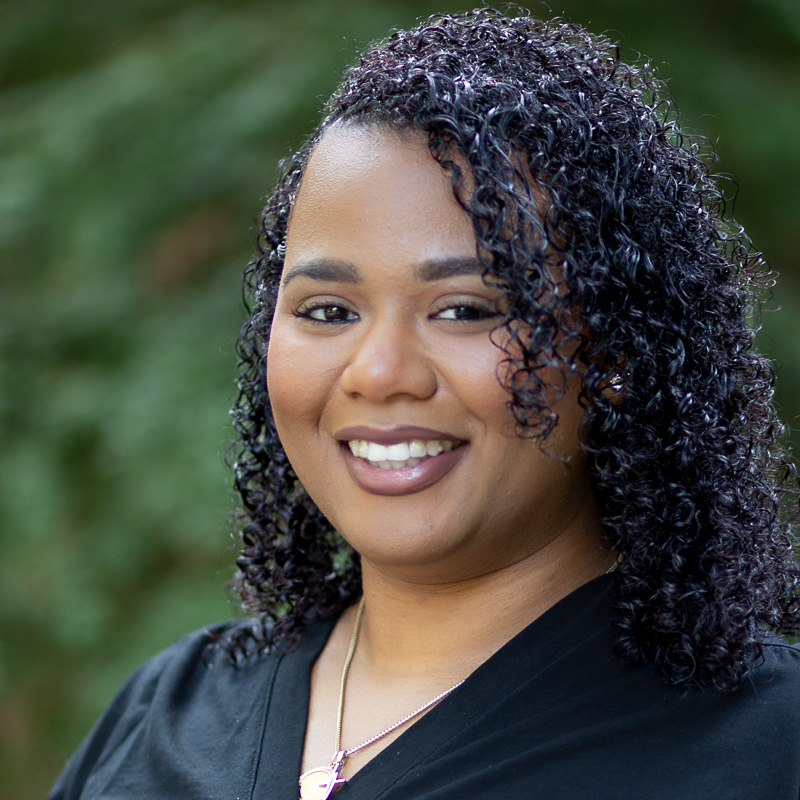 Headshot of Celine Osborne, with dark, curly shoulder-length hair, smiling showing teeth, wearing dark v-neck shirt and gold necklace, greenery in the background